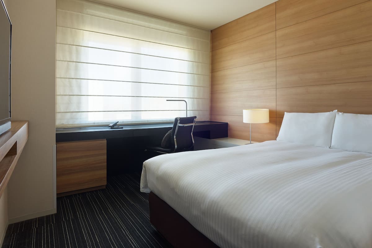 Quarto de hotel com cama, escrivaninha, cadeira, luminária e janela com persianas.