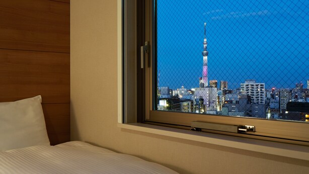 Une chambre d’hôtel dotée d’une grande fenêtre donnant sur une vue panoramique de la ville au crépuscule, avec une tour imposante en évidence.