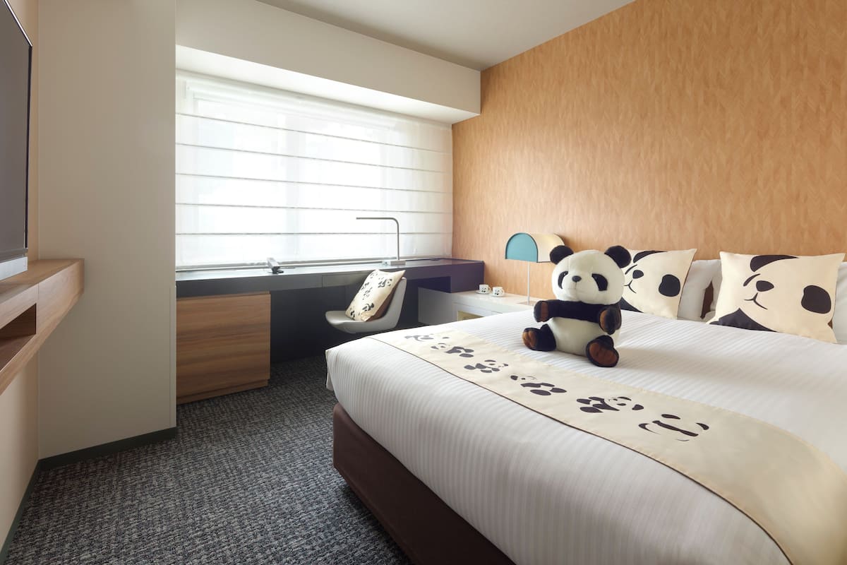 Quarto de hotel com cama, escrivaninha, cadeira, televisão, janela com persianas e almofada com tema de panda.