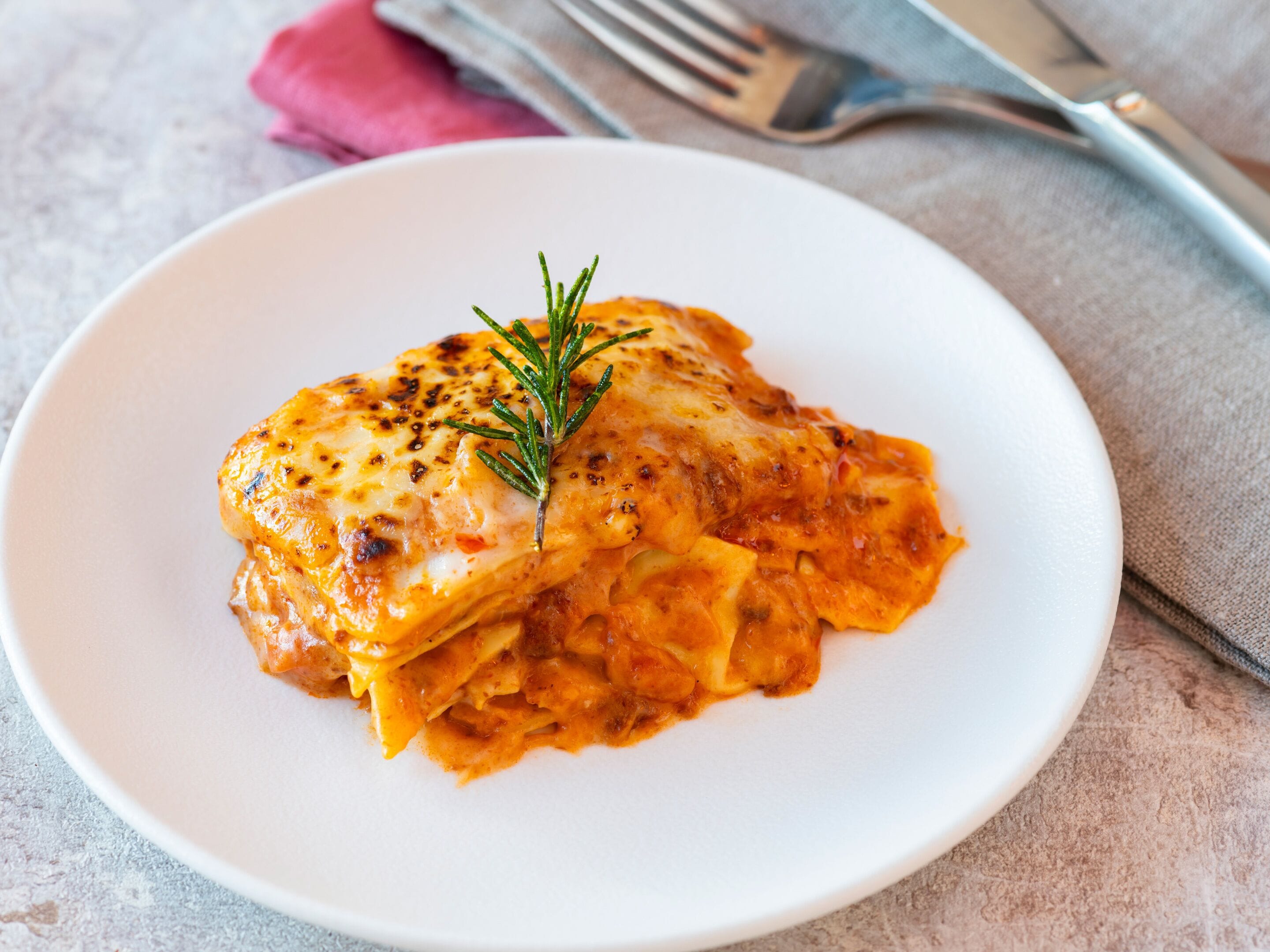 A plate of lasagna with a sprig of rosemary on top, served on a white plate.