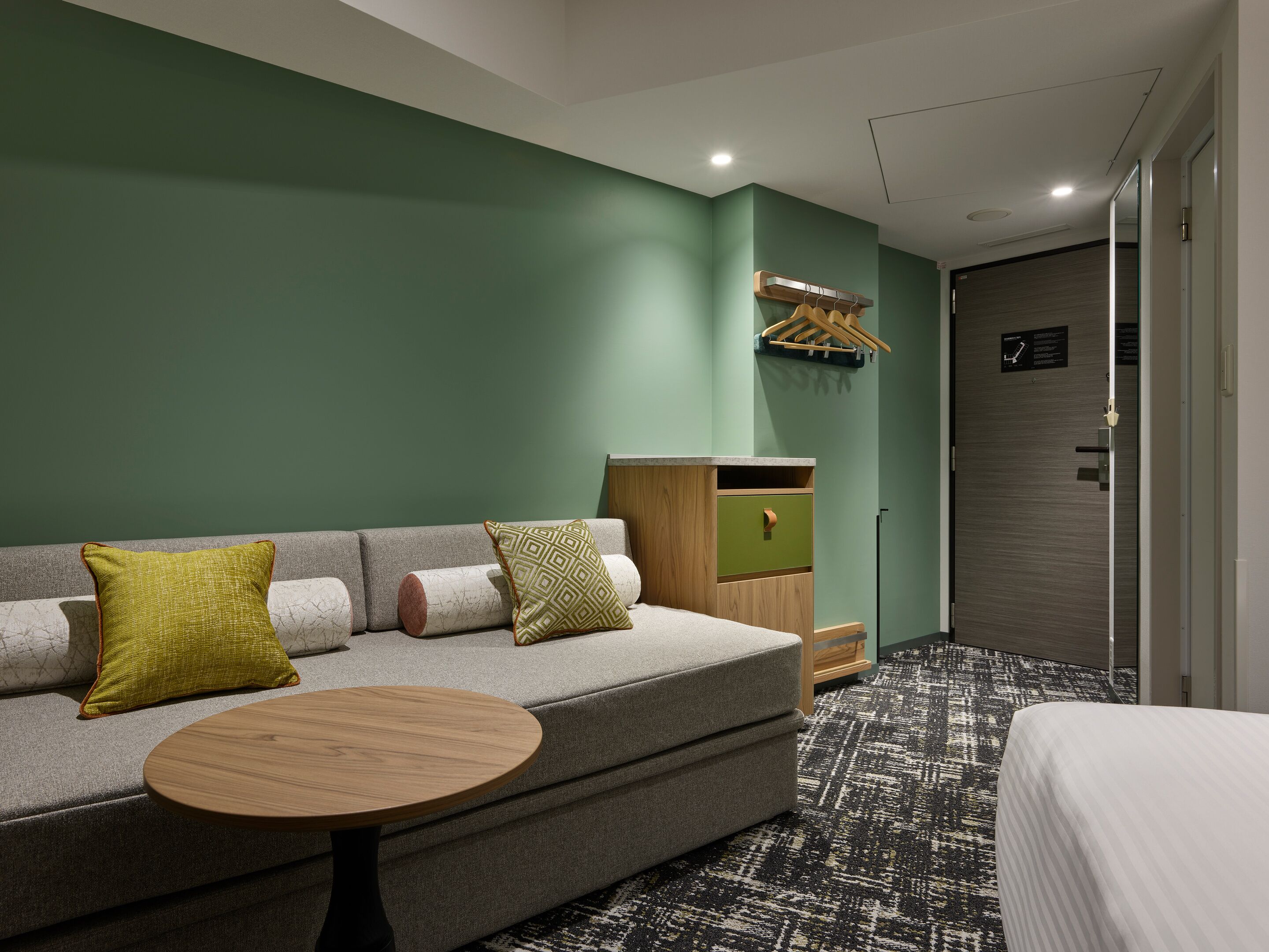 Una habitación de hotel moderna con un sofá gris, dos almohadones estampados, una mesa redonda de madera y una alfombra con diseño.