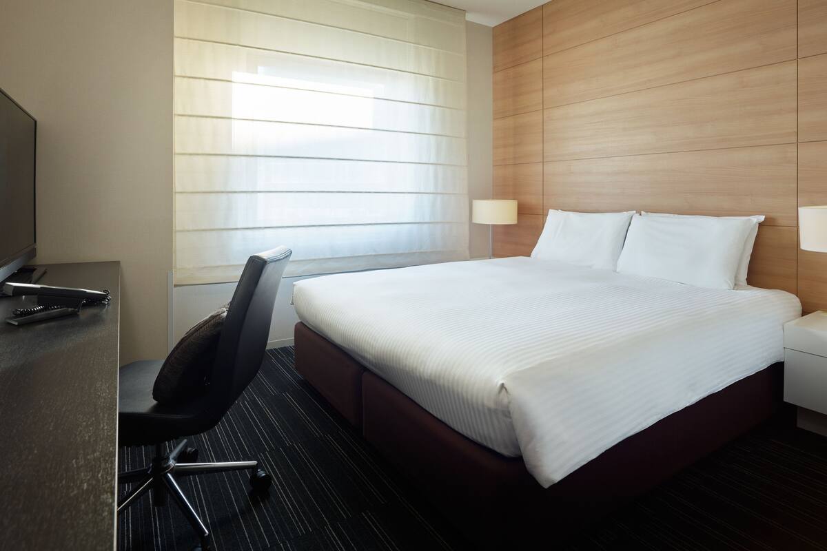 Quarto de hotel com cama, escrivaninha, cadeira, televisão e janela com persianas.