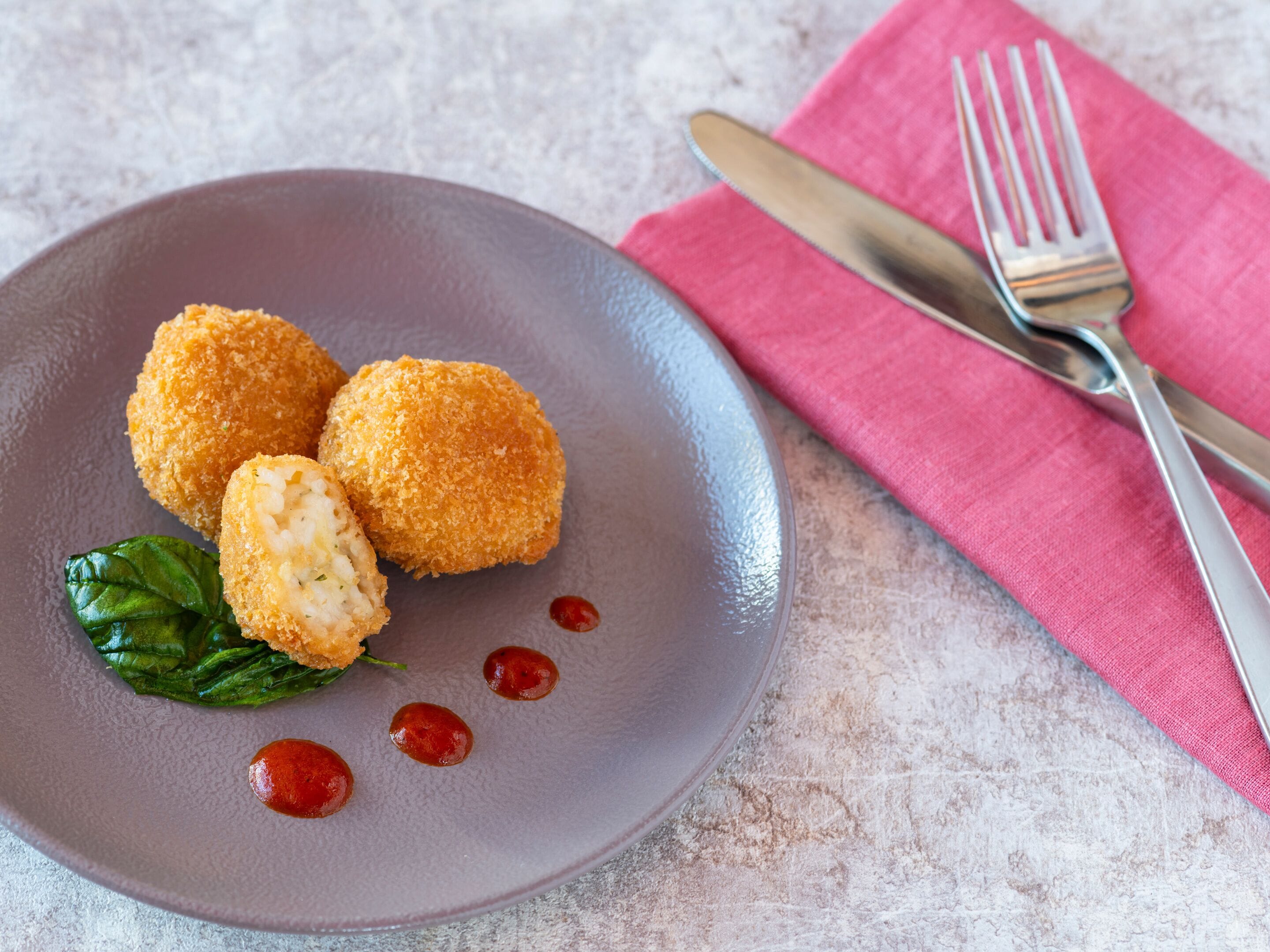 A plate with three golden-brown croquettes, a green leaf garnish, and a few red sauce dots on a textured grey plate.