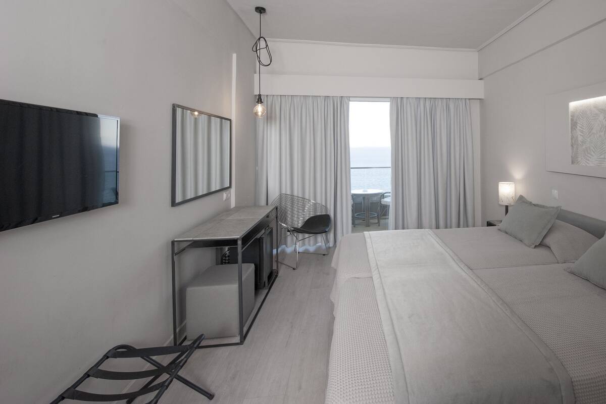 Habitación de hotel moderna con una cama grande, un escritorio, una silla, un televisor y vistas al mar tras las cortinas.