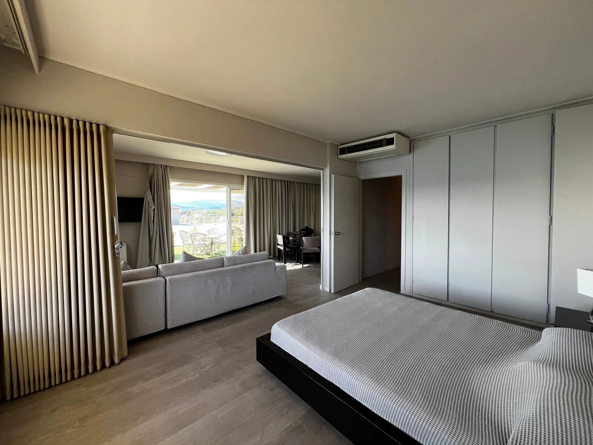 A modern hotel room with a large bed, a sofa, a dining area, and a view of the outdoors.