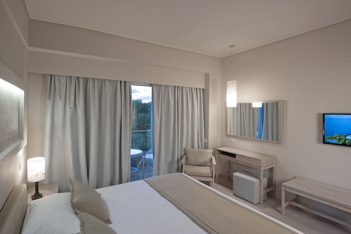 Habitación de hotel con cama, escritorio, silla, televisor y balcón con cortinas.