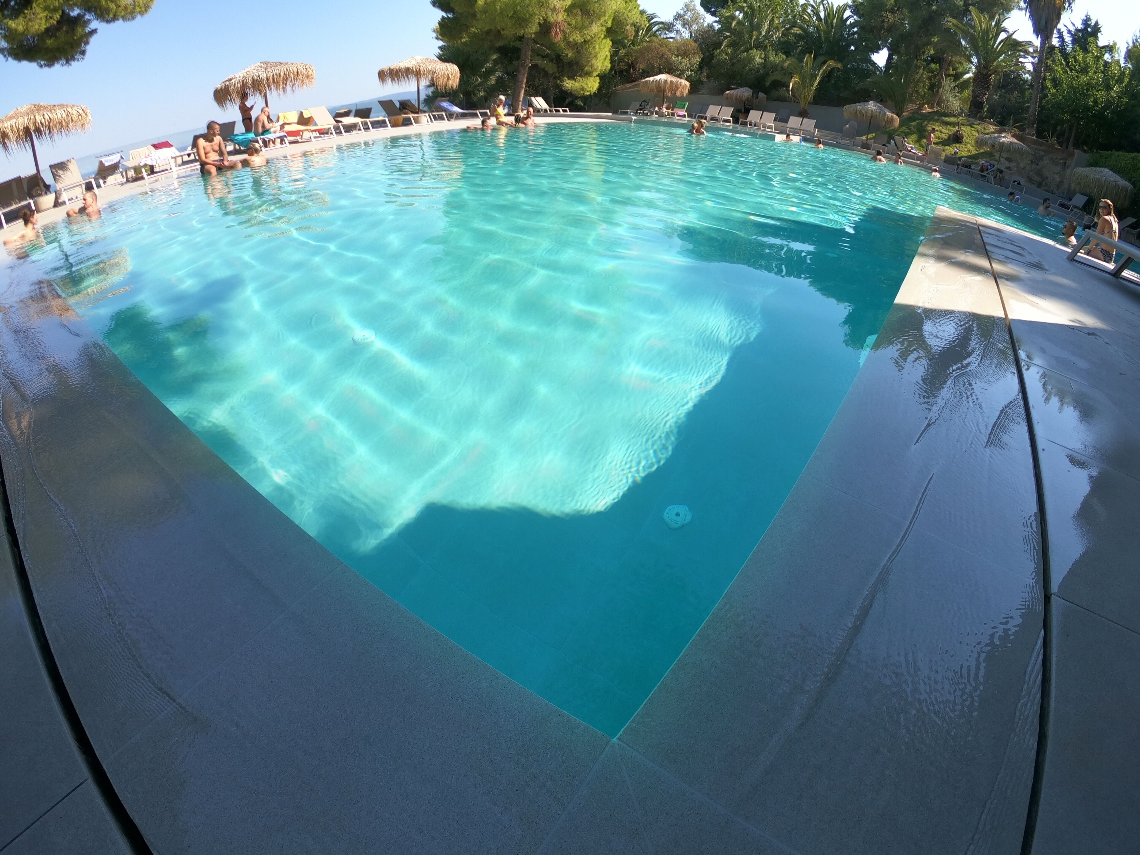 A clear blue pool with lounge chairs and umbrellas, surrounded by trees and a view of the sea.