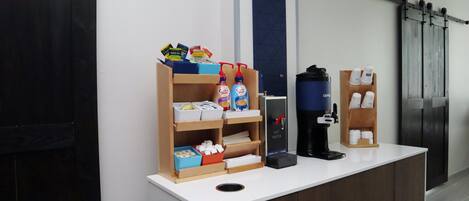 Zona de recepción de un hotel con mostrador, máquina de café y estantes con aperitivos y artículos de tocador.