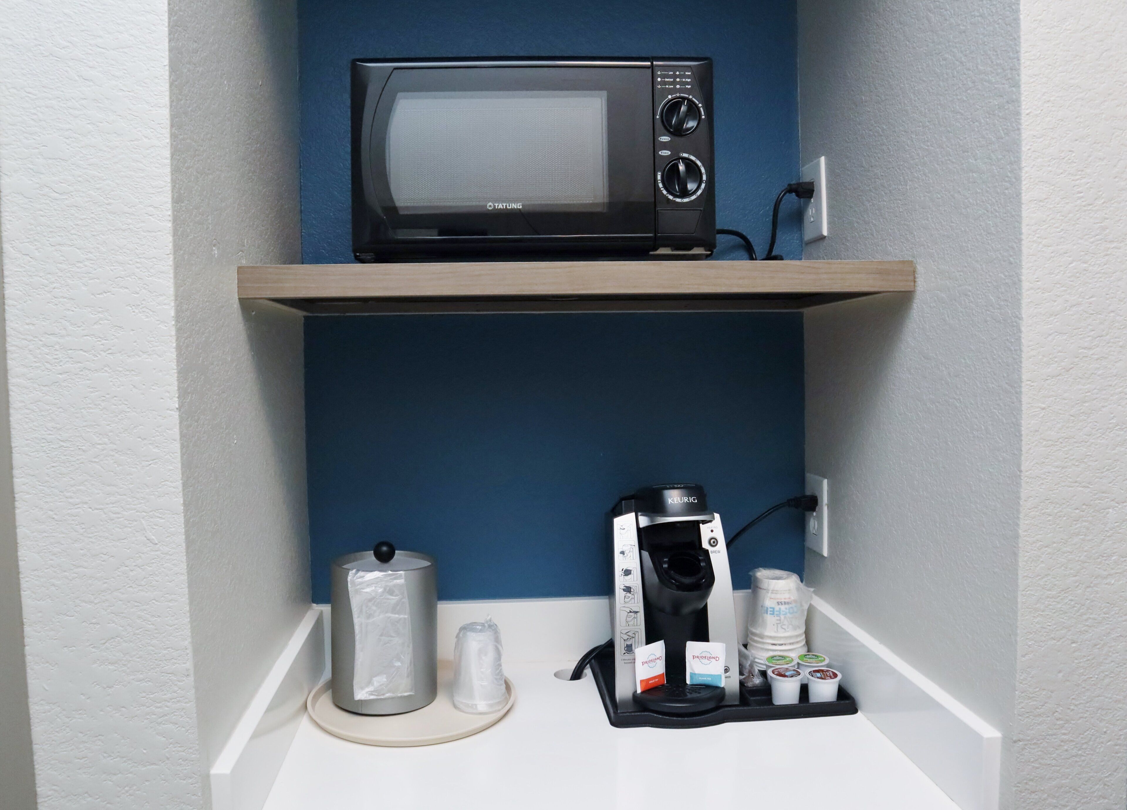 A kitchenette with a microwave on a shelf, a Keurig coffee maker, a coffee pod holder, and a water dispenser.