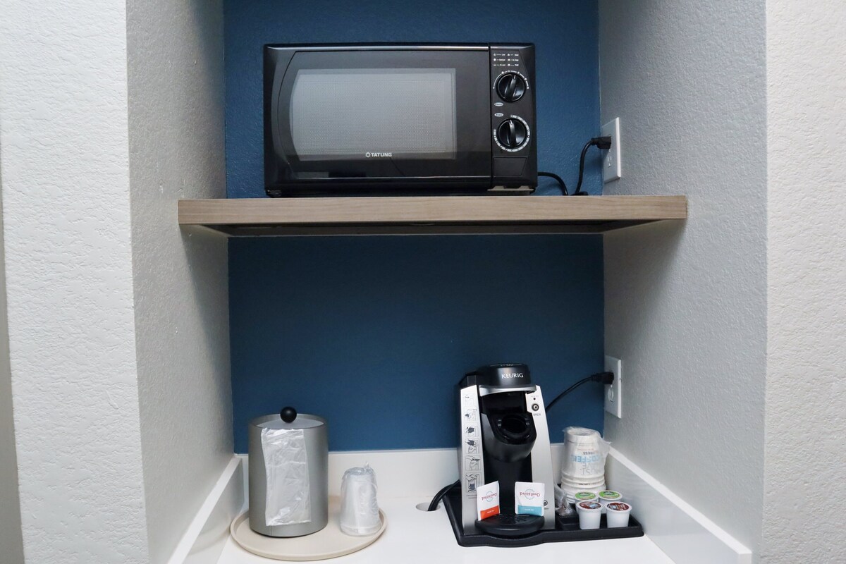 Et tekøkken med en mikroovn på en hylde, en Keurig kaffemaskine, en holder til kaffekapsler og en vanddispenser.