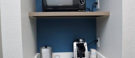A kitchenette with a microwave on a shelf, a Keurig coffee maker, a coffee pod holder, and a water dispenser.