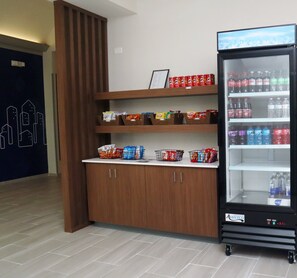 En automtomat fyldt med drikkevarer og snacks, et træskab med kurve til snacks, og en dør med et bybillede som mural.