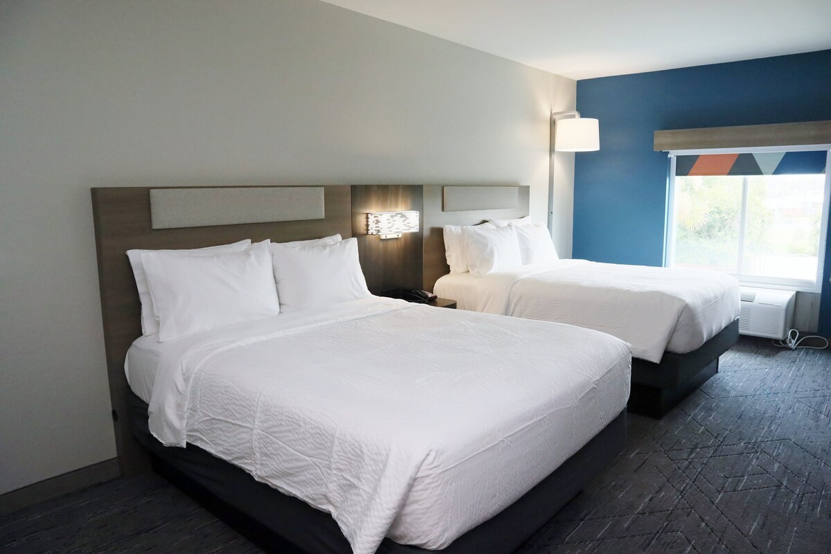 A hotel room with two beds, a nightstand, a wall-mounted lamp, and a window with blinds.