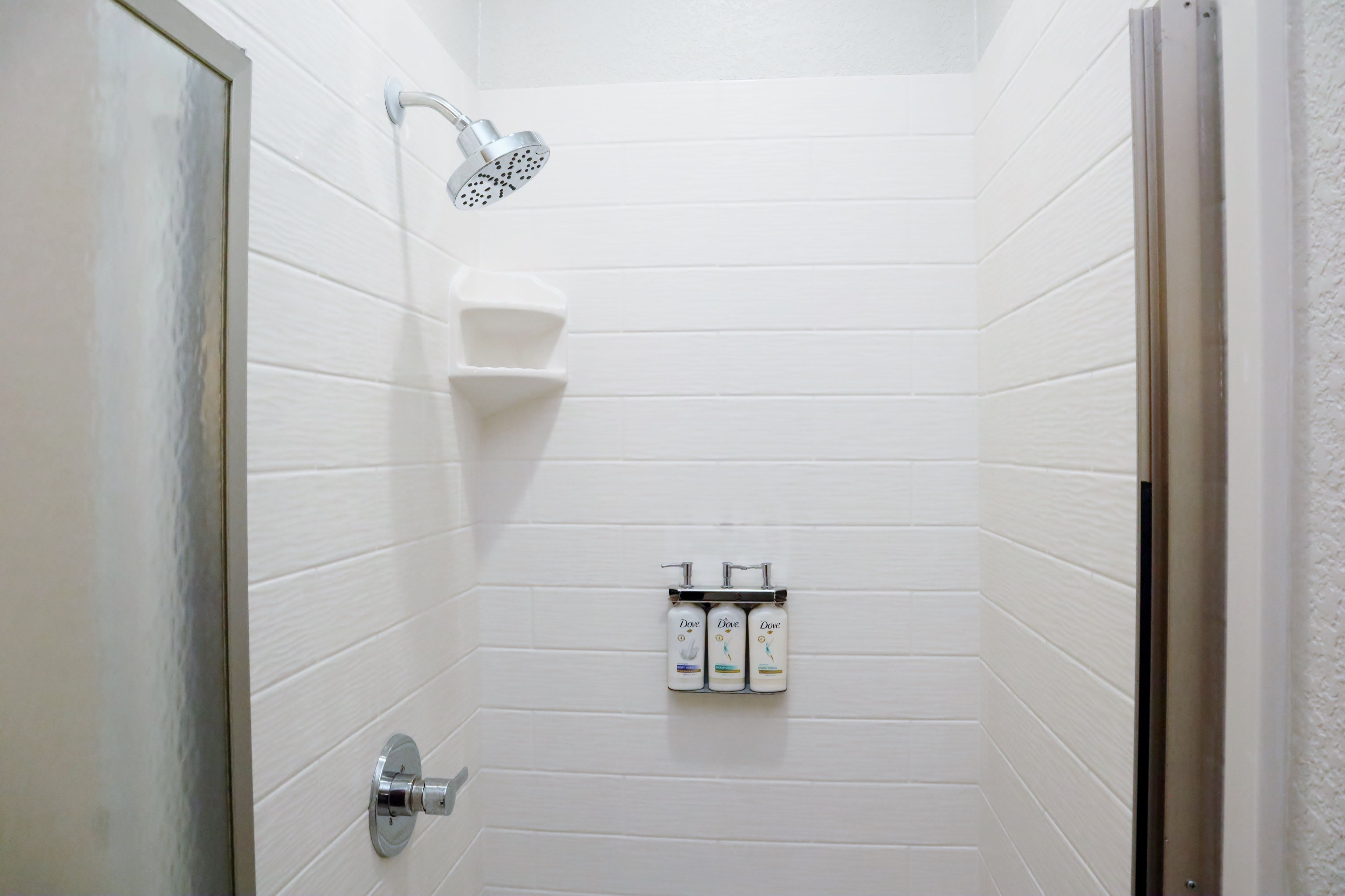 A bathroom with white tiled walls, a showerhead, and a soap dispenser.
