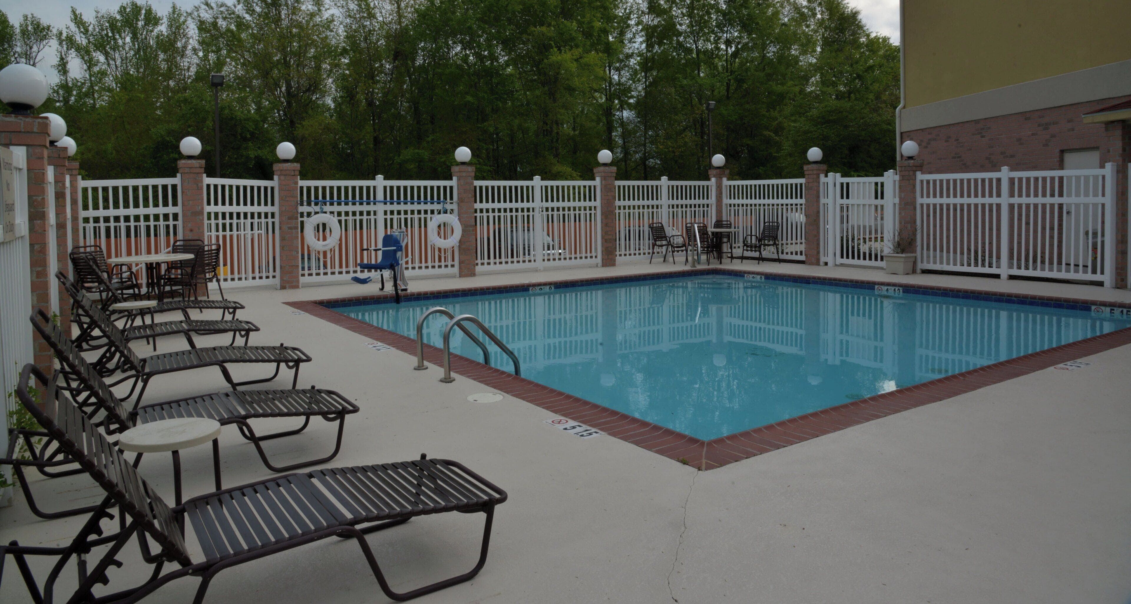 A swimming pool with a white fence, lounge chairs, and a brick wall.