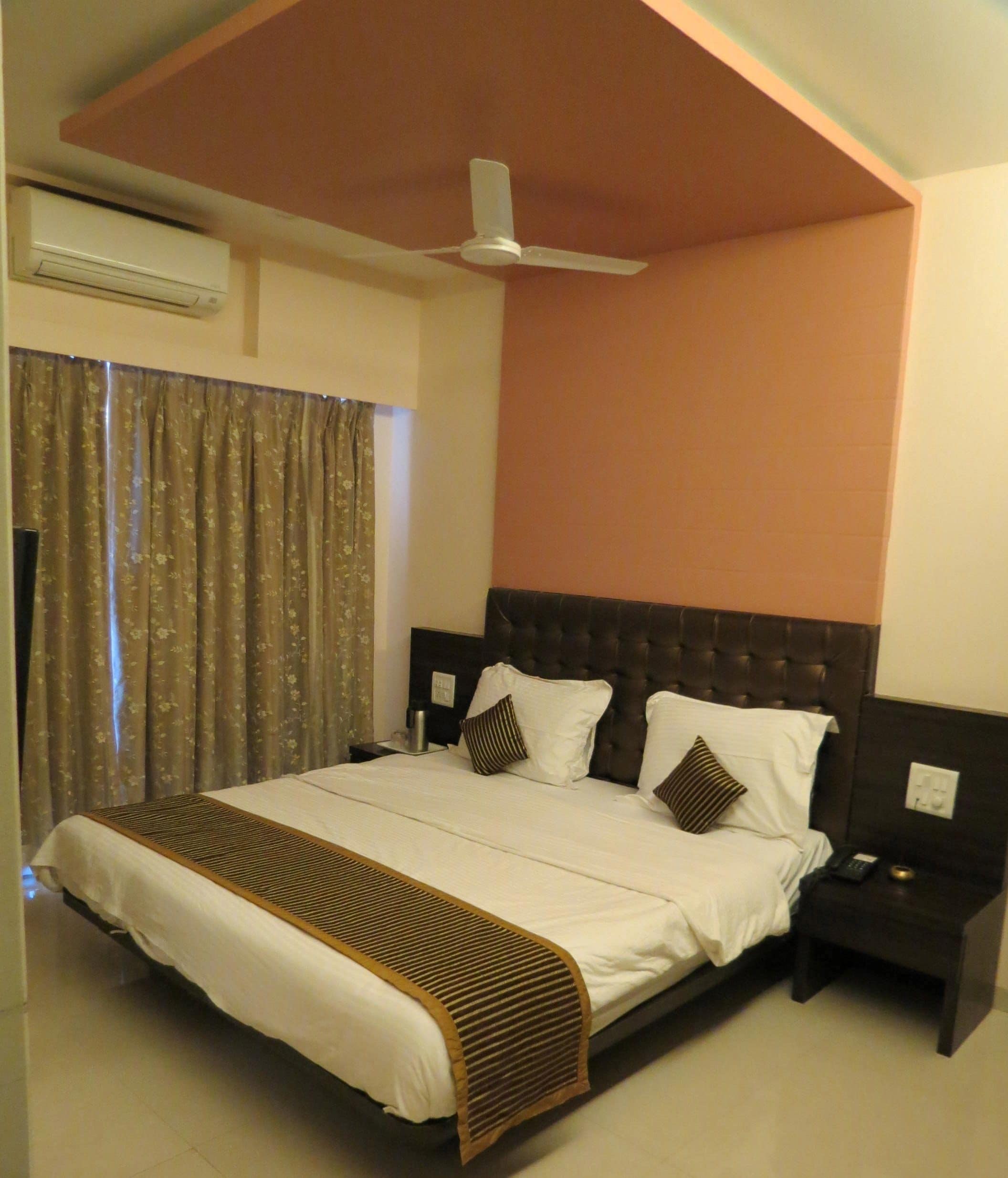 A bedroom with a large bed, a ceiling fan, an air conditioning unit, a nightstand, and a window with curtains.