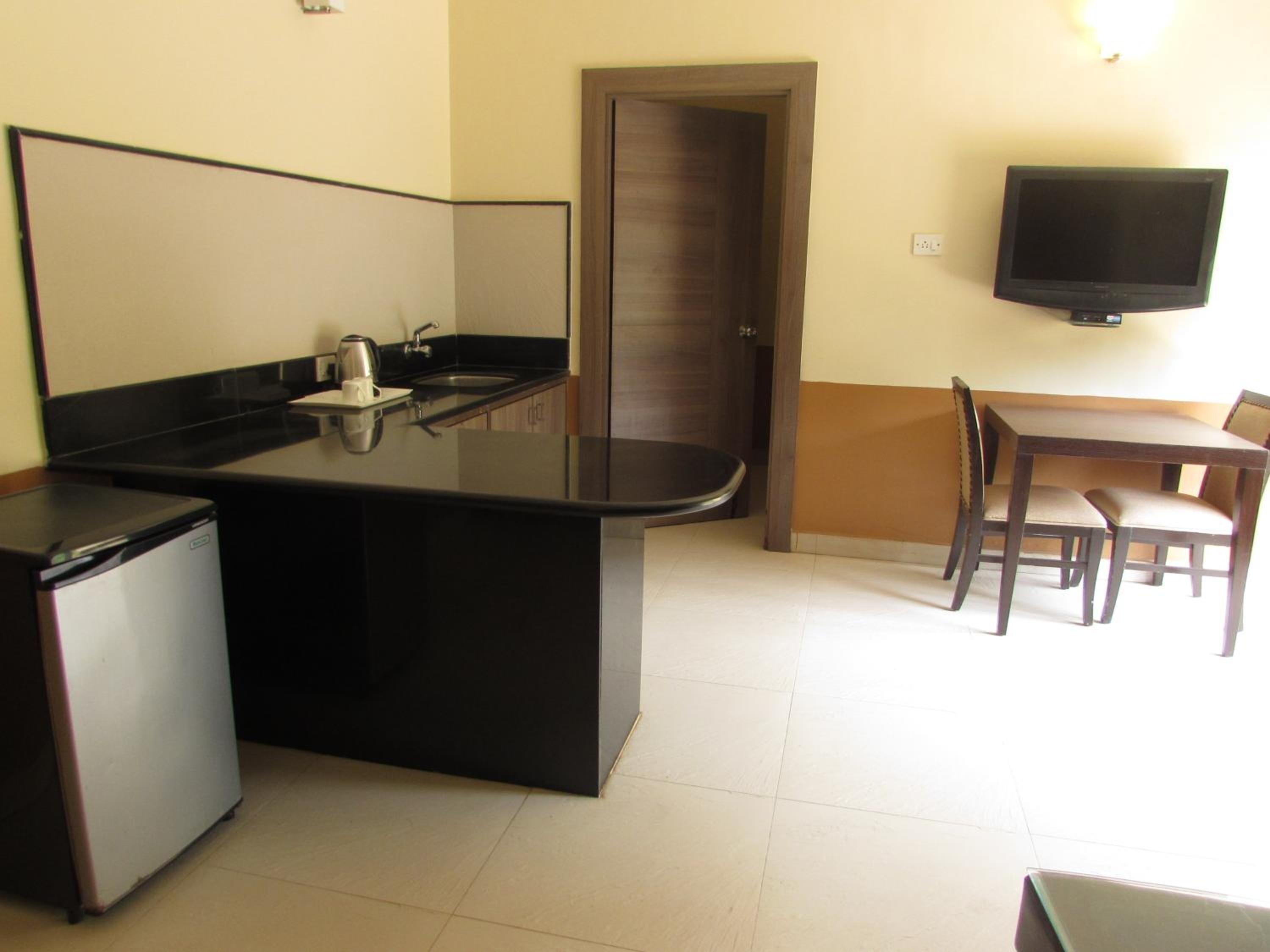 A compact kitchen with a refrigerator, a coffee maker, and a sink. There is a dining table with chairs and a flat-screen TV mounted on the wall.