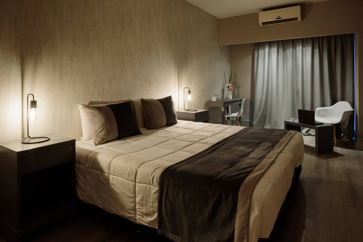 A modern hotel room with a large bed, bedside tables with lamps, a desk with a chair, and a large window with curtains.