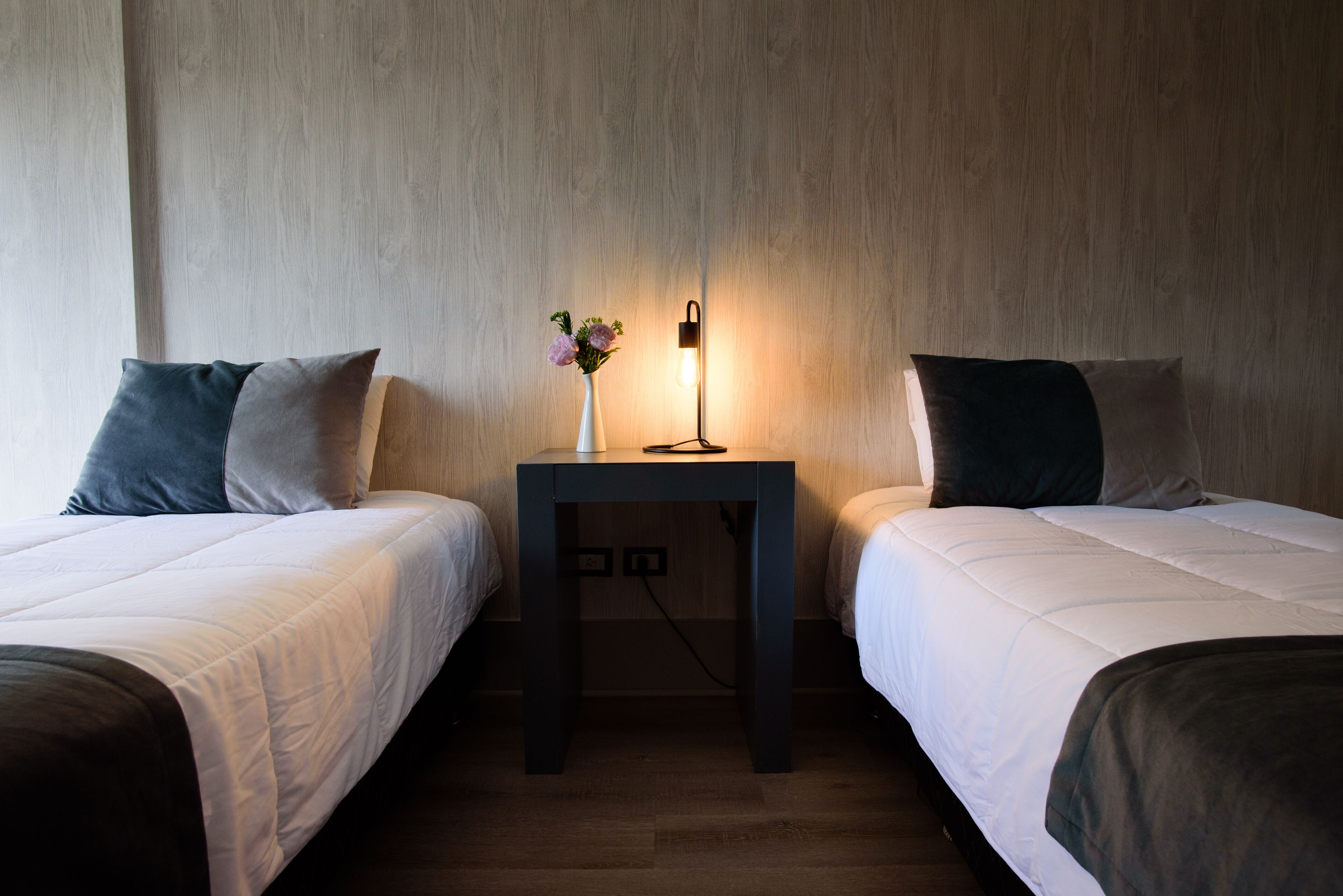 A hotel room with two single beds, a small table with a vase of flowers, and a wall-mounted light.