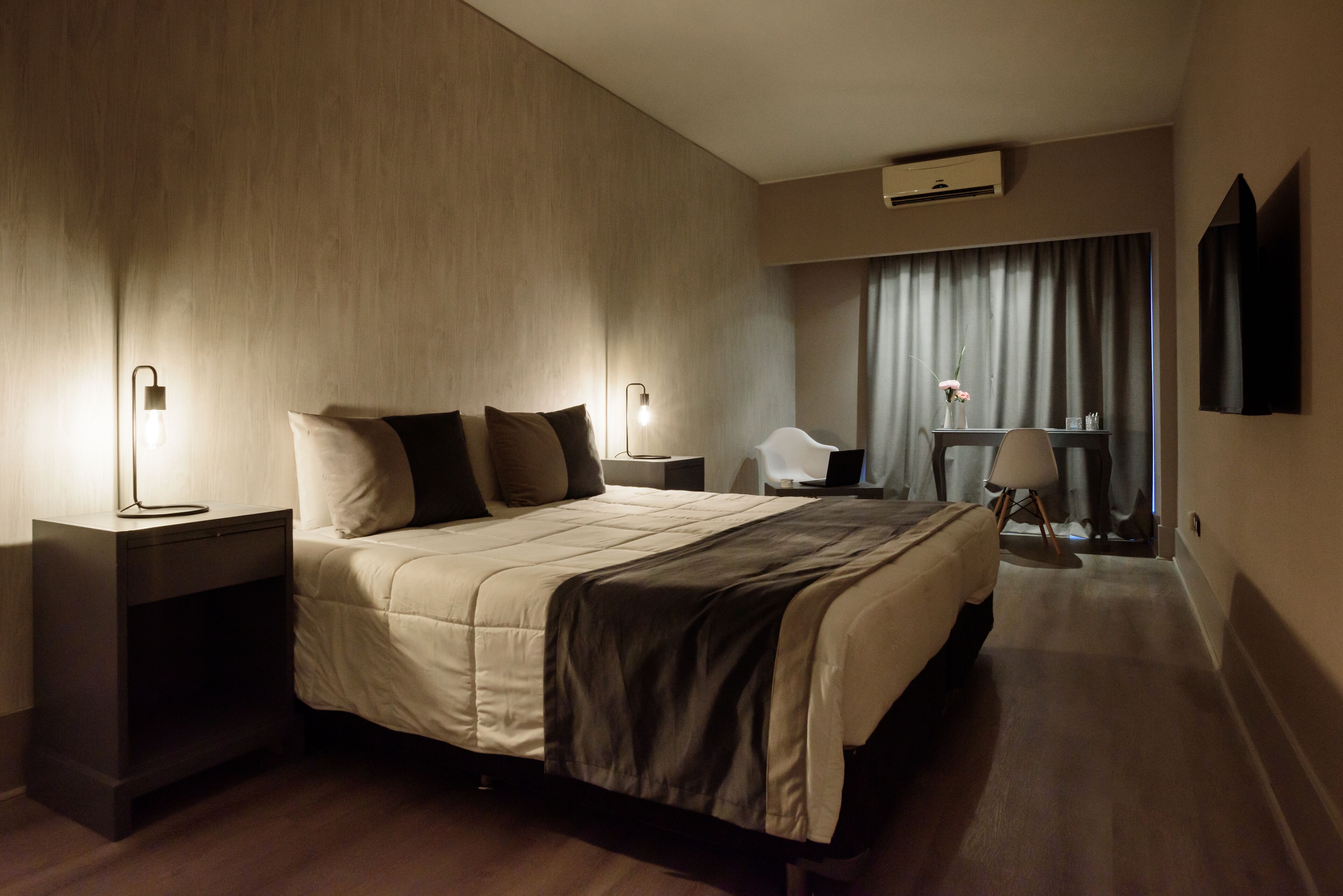 A modern hotel room with a large bed, bedside tables with lamps, a desk with a chair, and a large window with curtains.