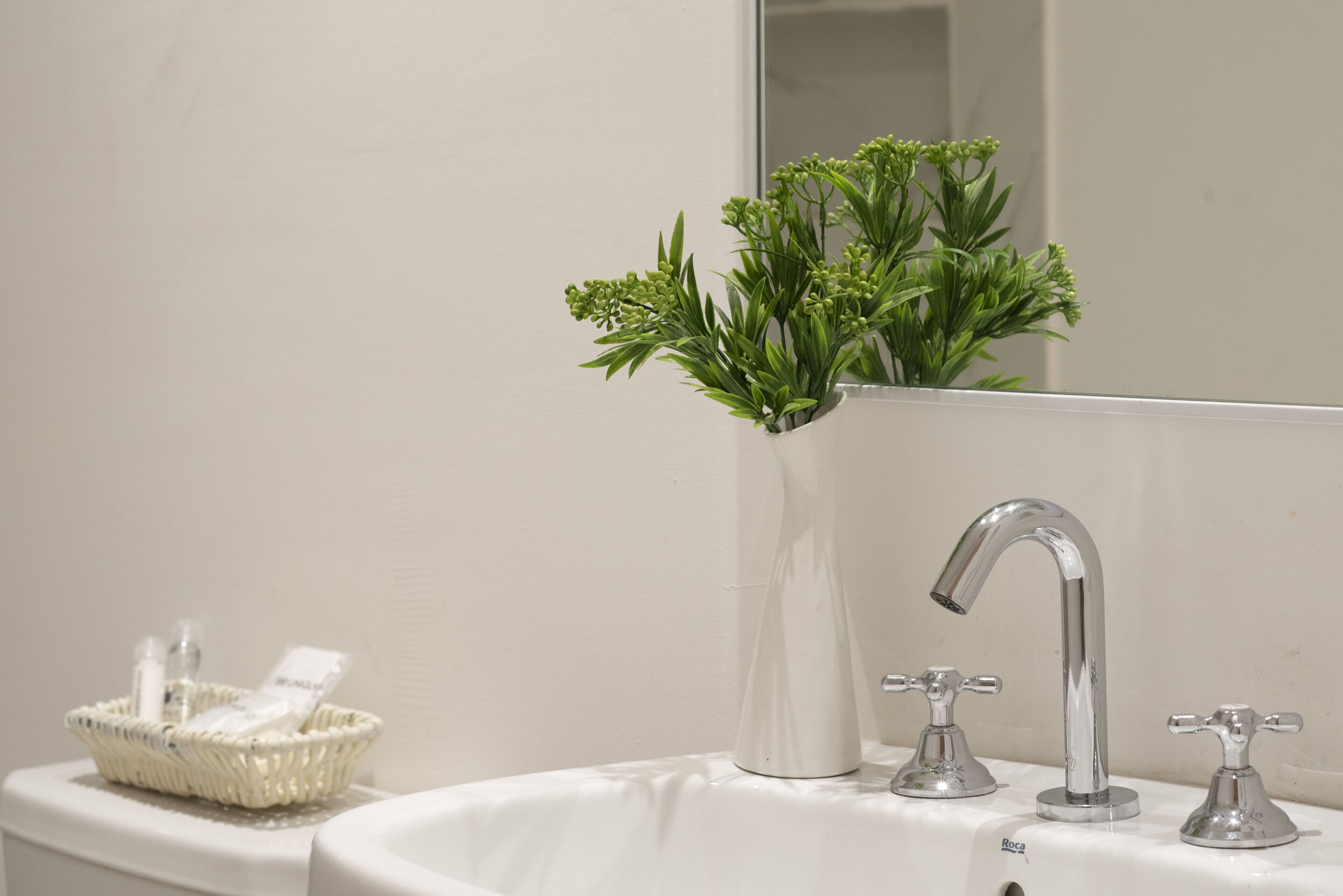 A bathroom with a white sink, two faucets, a mirror, a white vase with green plants, and a basket with toiletries.