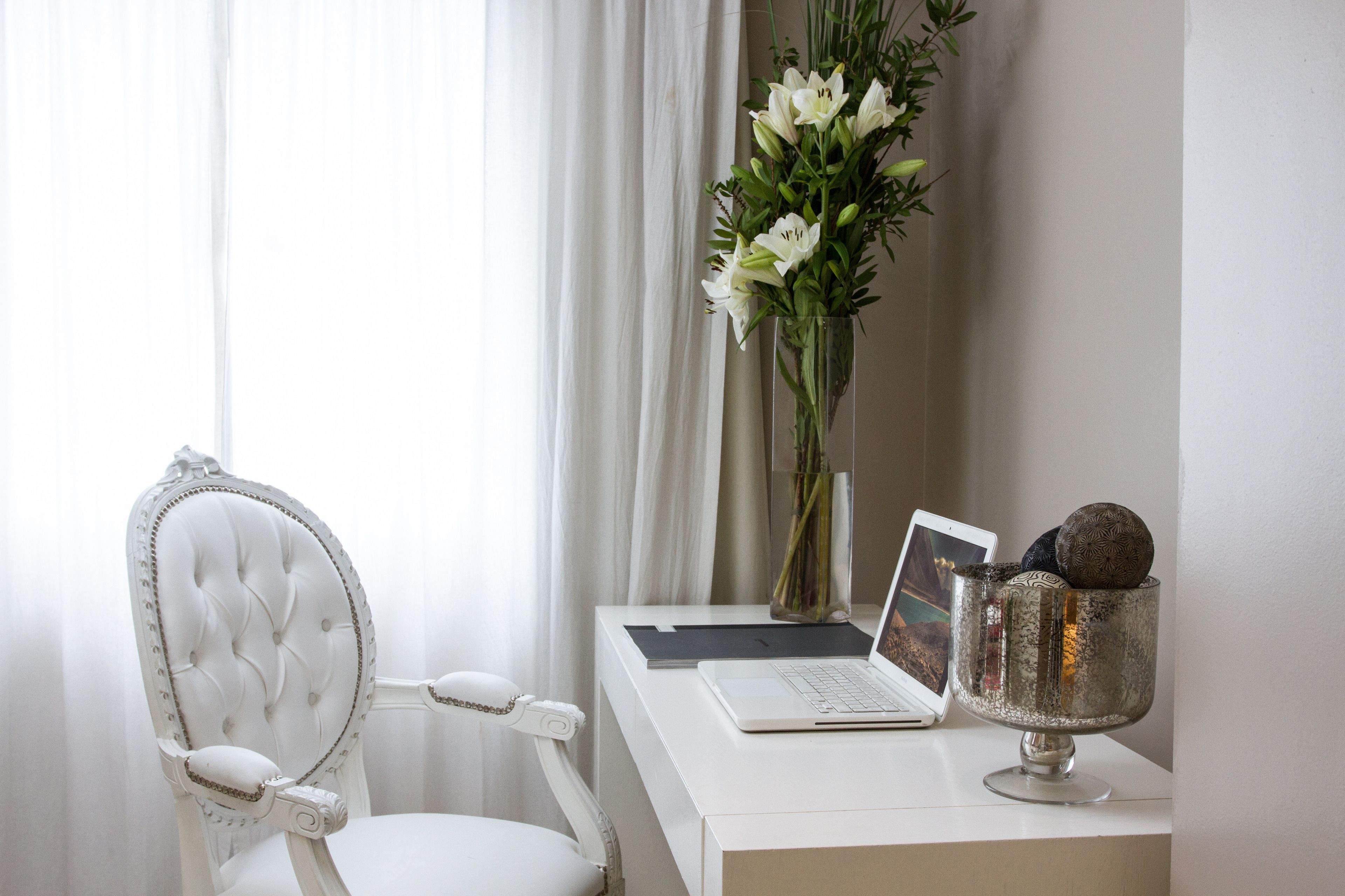 Een witte stoel met een gestuufde rugleuning, een laptop op een wit bureau, een vaas met witte bloemen, en een decoratieve metalen kom met een wereldbol en een boek.
