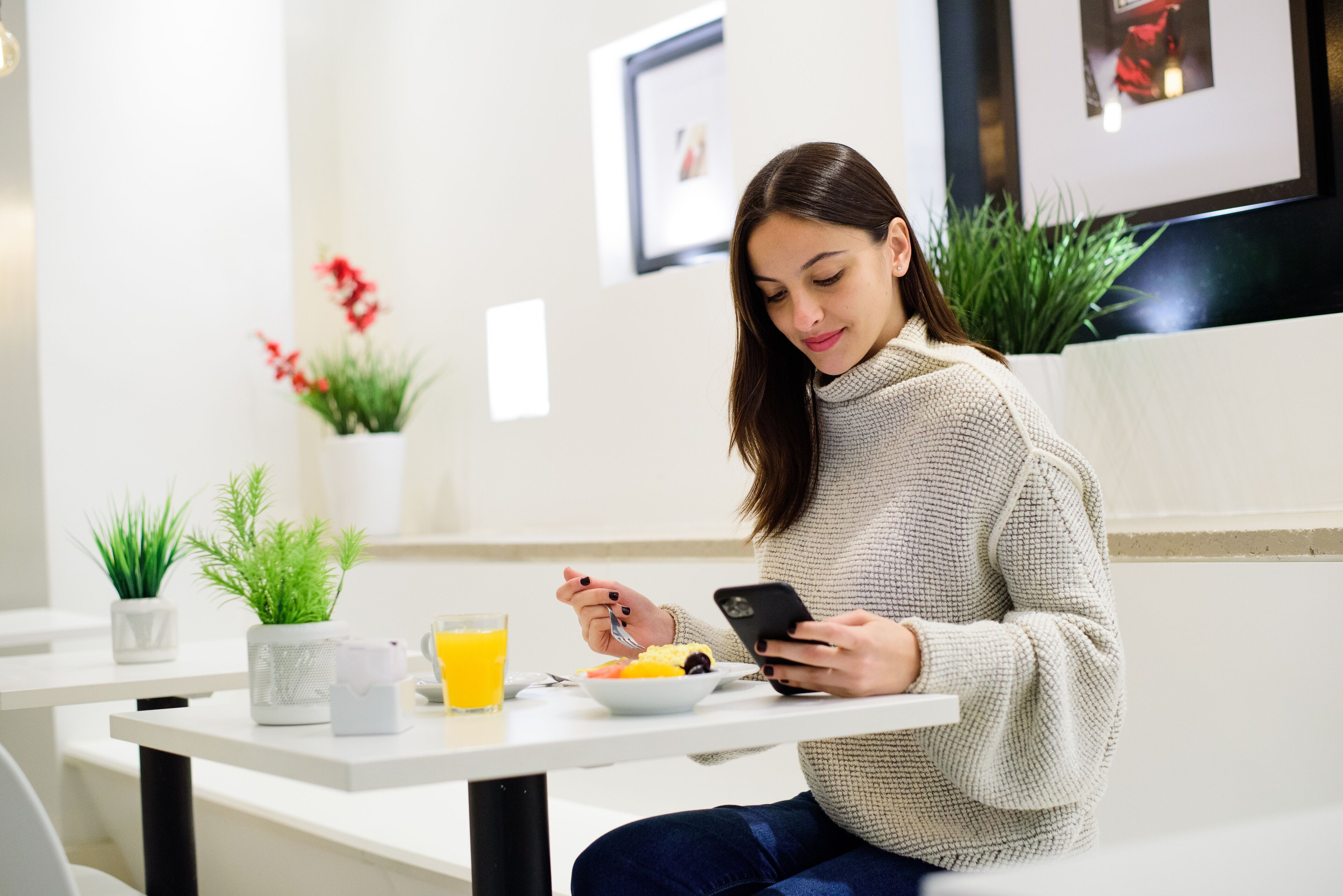 A person sitting at a table with a bowl of food, a glass of orange juice, and a smartphone. There are potted plants on the table and a framed picture on the wall behind.