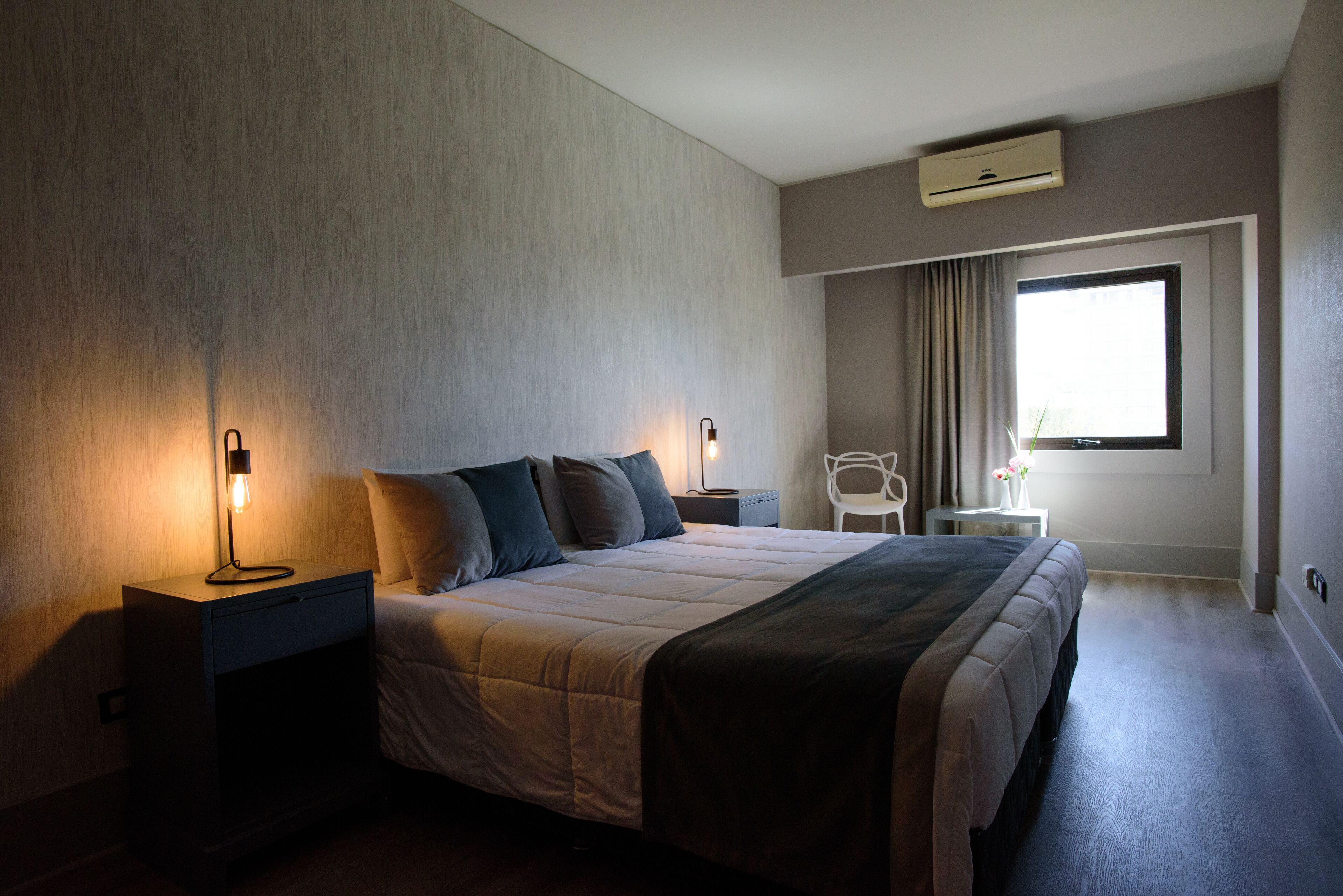A modern hotel room with a large bed, bedside tables, a nightstand, a chair, a small table with a vase, and a window with curtains.