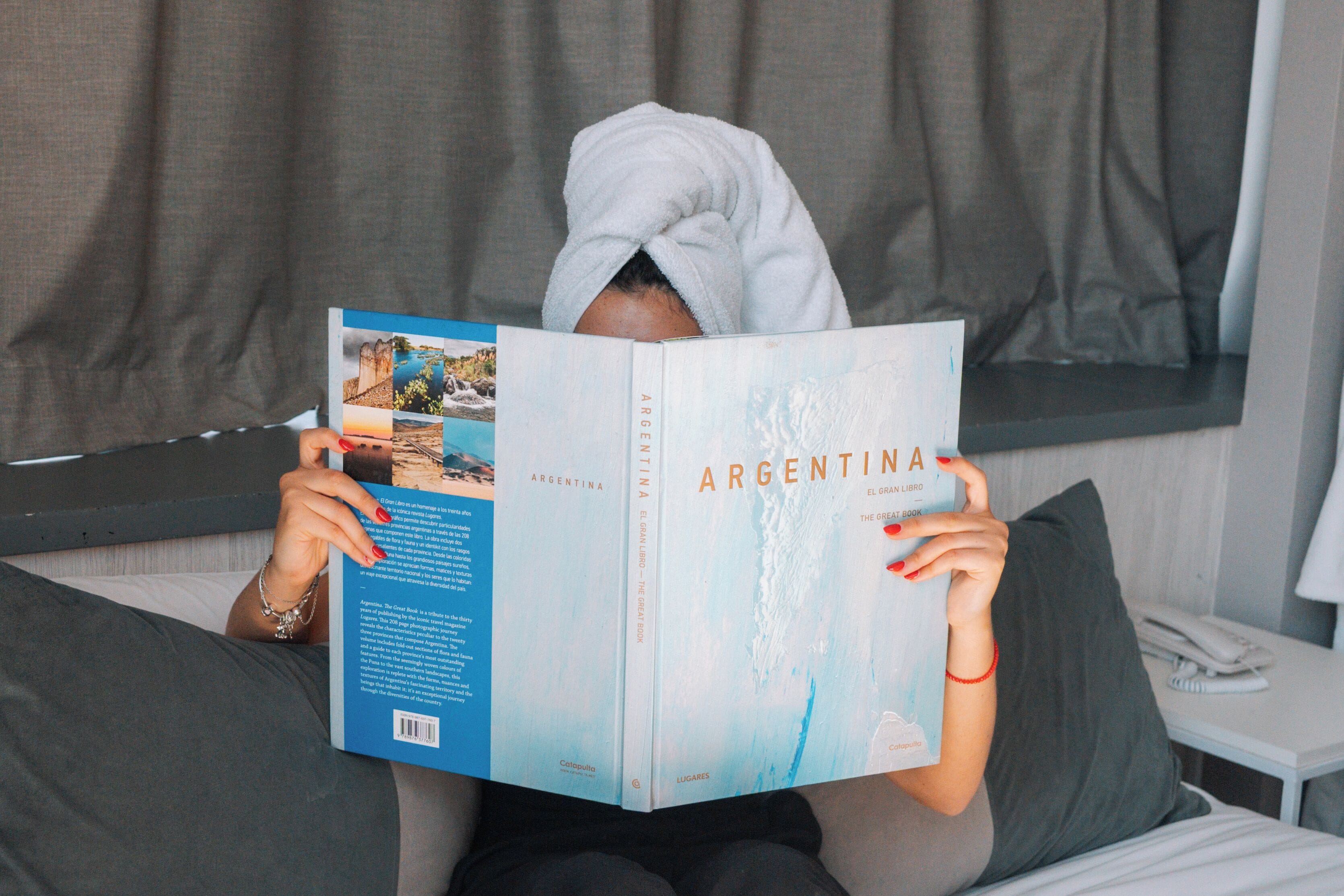 A person reading a book titled 'Argentina' in a room with a bed, pillows, and a small table with a lamp.