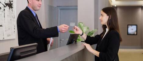 A reception area with a reception desk, a man handing a card to a woman, and a plant in the background.