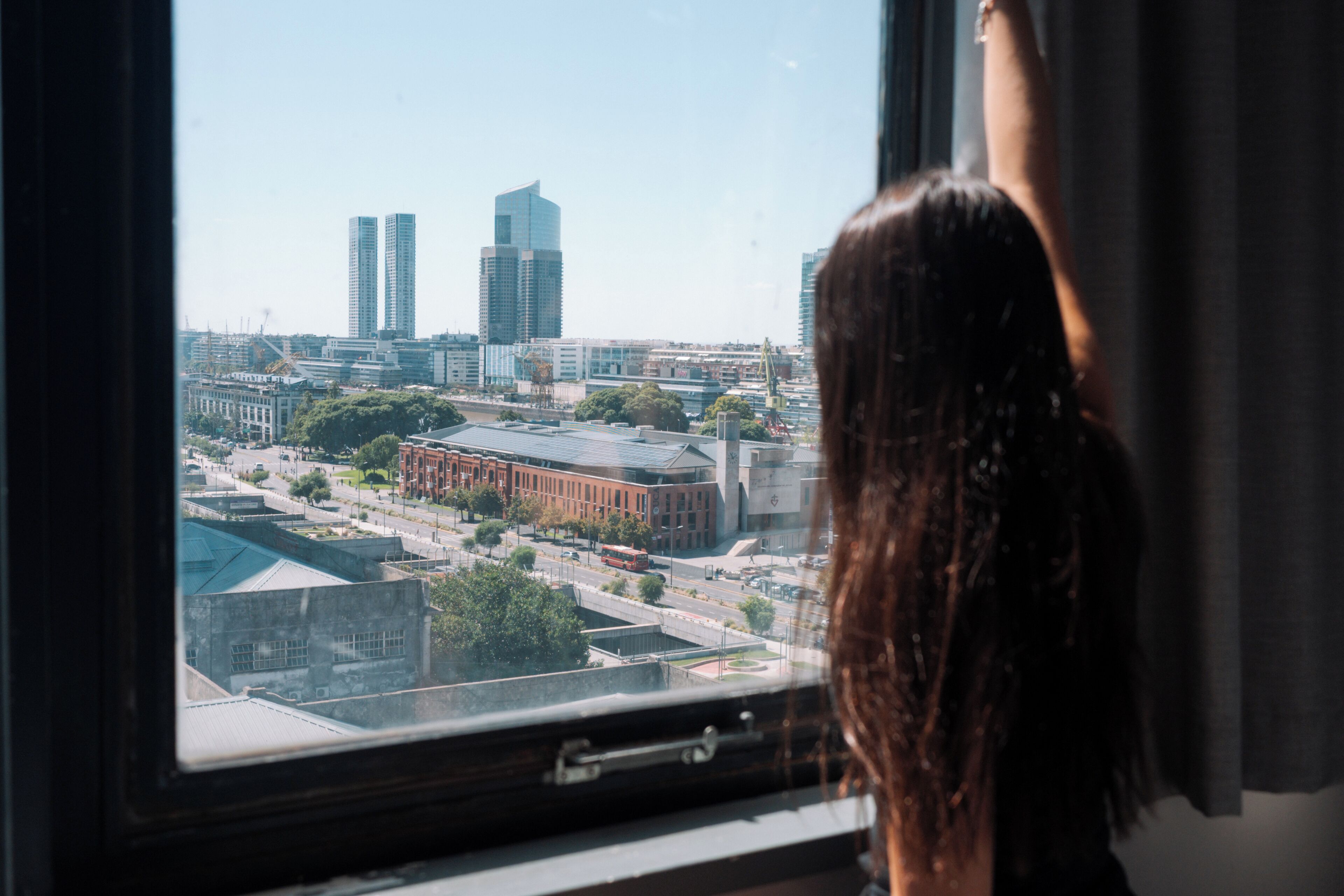 A person looking out of a window at a cityscape with buildings and a road.