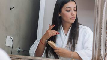 Una persona vestida con una bata blanca de pie frente a un espejo en un baño con pared beige, una puerta blanca y un espejo enmarcado.