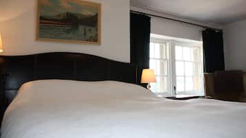 A large bed with a headboard, a painting on the wall, a window with curtains, and a chair.