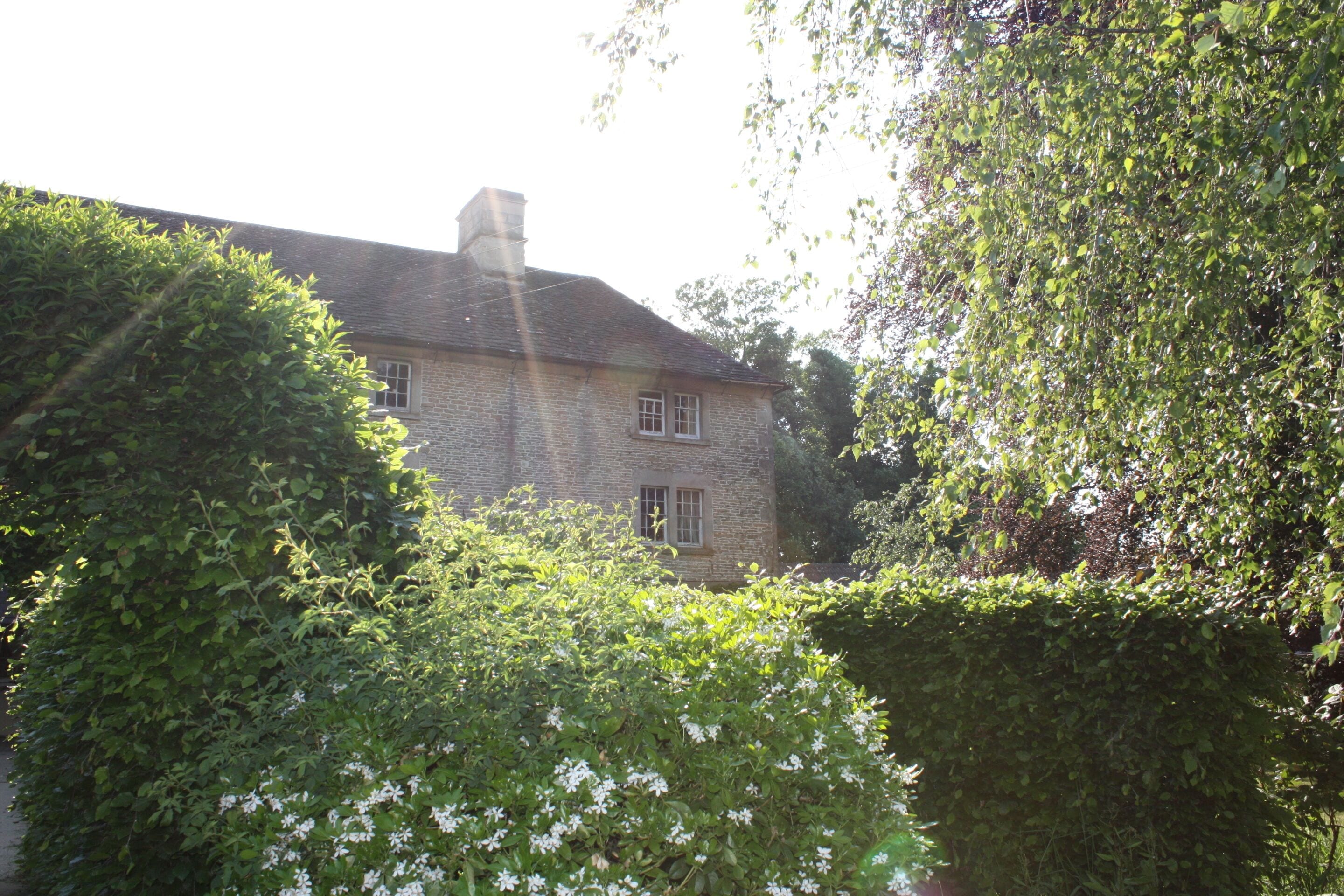 A two-story stone house with a chimney, surrounded by lush green foliage and flowering plants.