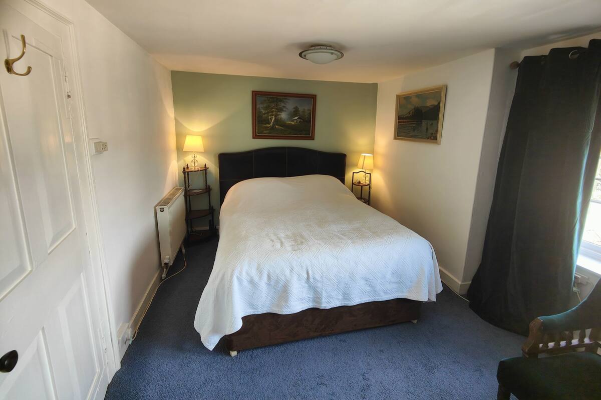 A neatly arranged bedroom with a large bed, two bedside lamps, a radiator, and two paintings on the wall.