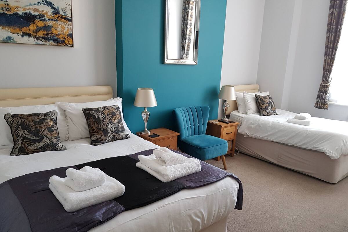 Habitación de hotel con dos camas, un sillón azul y un cuadro en la pared.