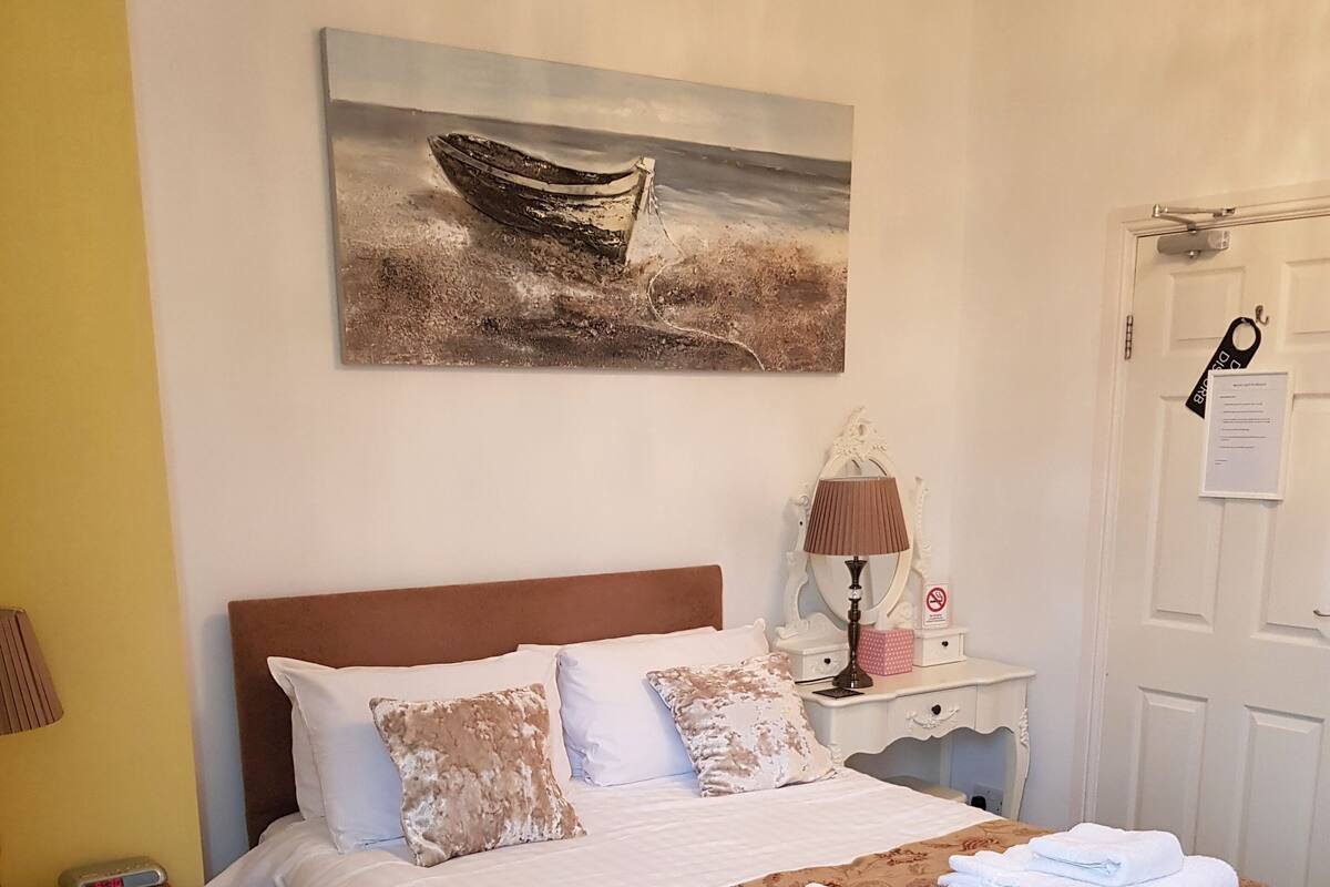 Una cama bien hecha con sábanas blancas y una colcha estampada con motivos florales, una mesita de noche con una lámpara y un pequeño adorno, y un cuadro de un barco en la pared.
