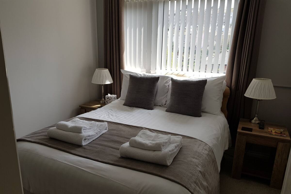 Una cama bien hecha con sábanas blancas y dos almohadas, una mesita de noche con lámpara y una ventana con persianas.