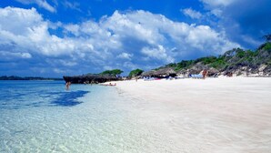 A beach with clear shallow waters, a sandy shore, and a few structures in the background. - Temple Point Resort (Watamu)