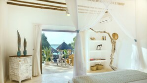 A bedroom with a canopy bed, a rustic side table, and a view of the beach and thatched-roof structures. - Temple Point Resort (Watamu)