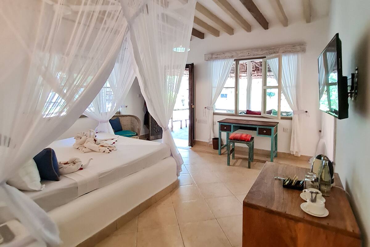 Una habitación con una cama bajo un dosel, una mesa de madera y un espejo, y un ventanal con cortinas.