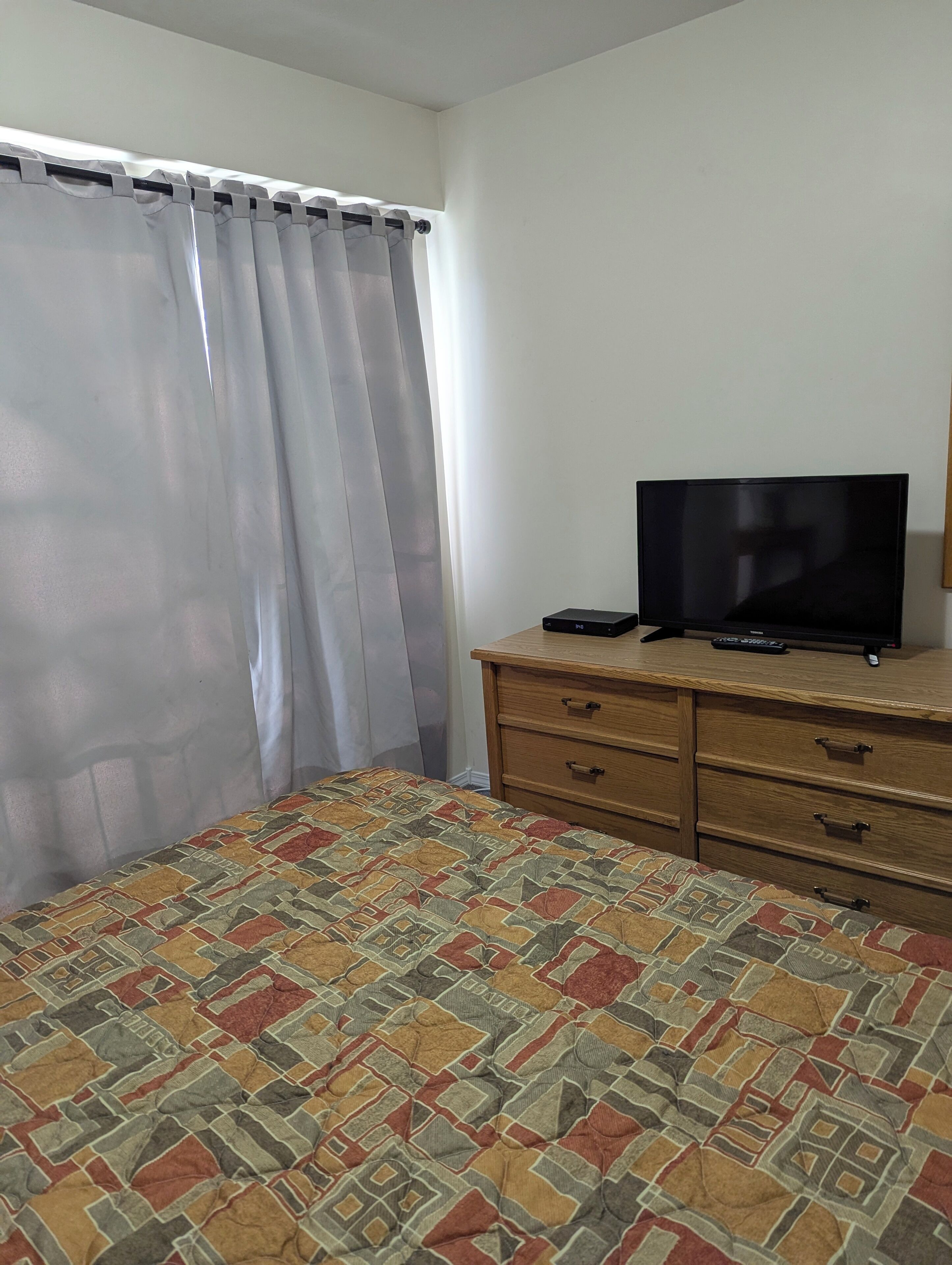 A bedroom with a bed, a TV on a wooden dresser, and a window with curtains.