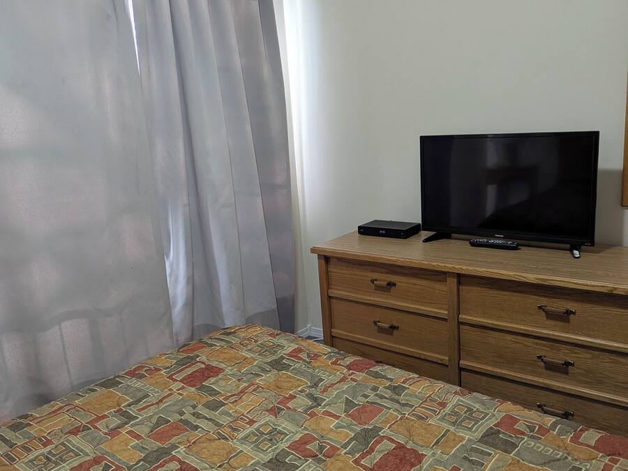 A bedroom with a bed, a TV on a wooden dresser, and a window with curtains.