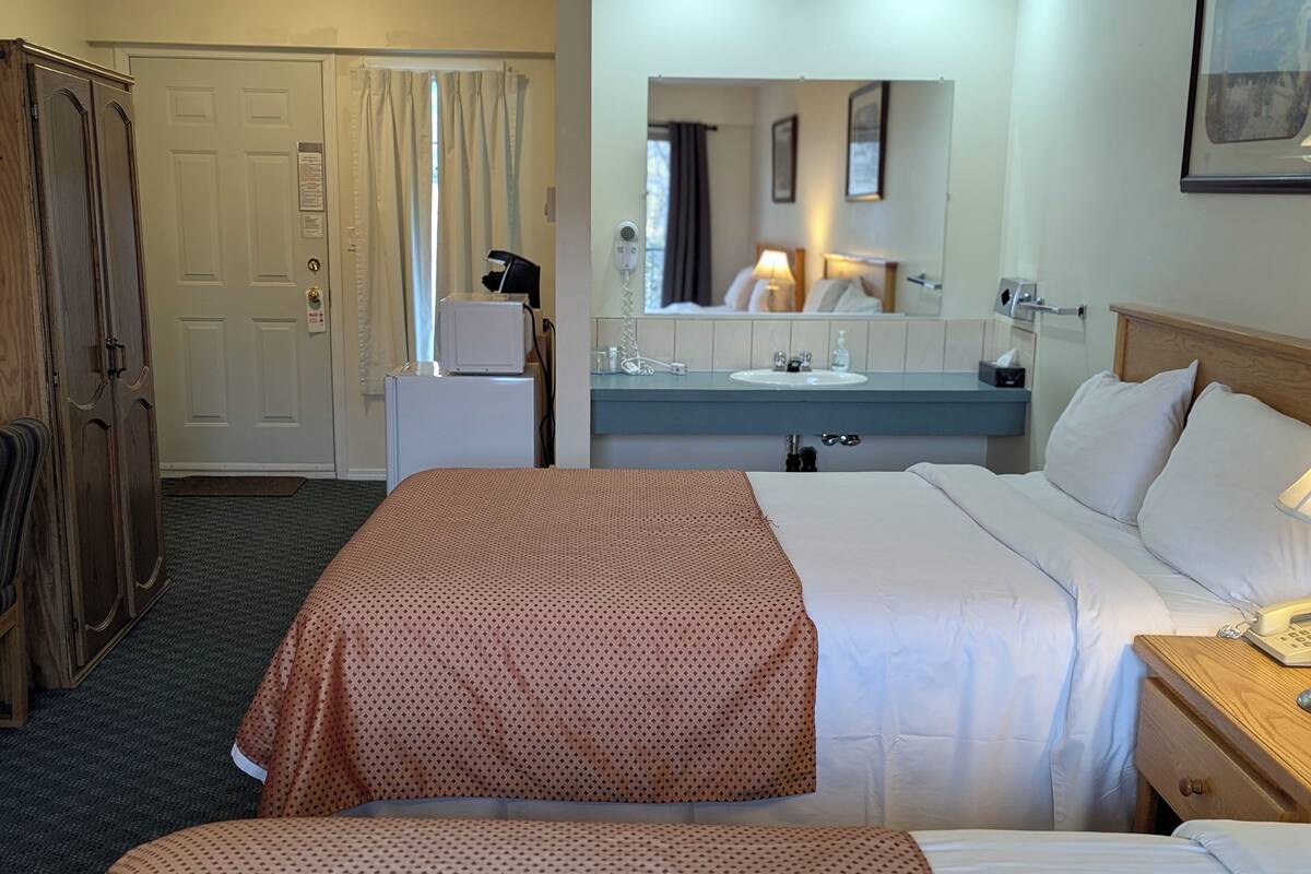 A hotel room with two beds, a desk, a mirror, a small refrigerator, and a wardrobe.