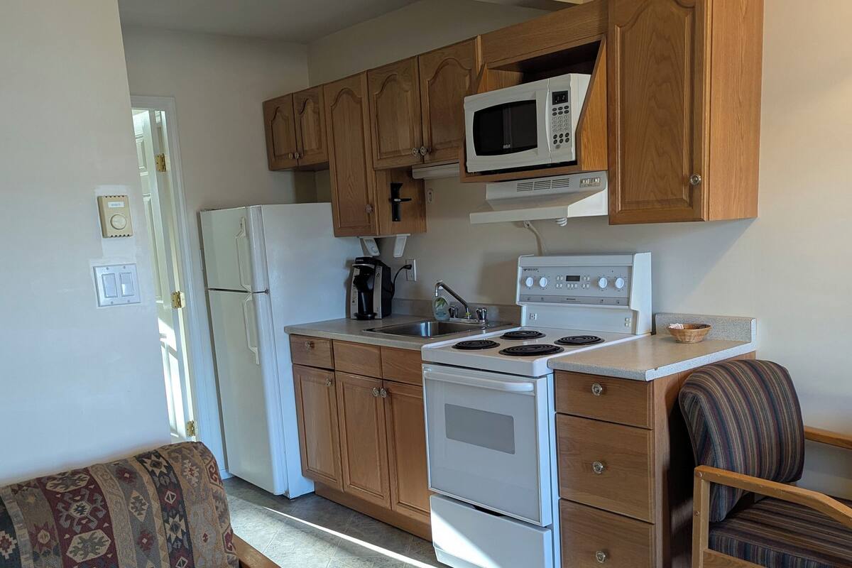 A compact kitchen with wooden cabinets, a microwave, a refrigerator, and a stove. There is a patterned sofa and a chair in the living area.