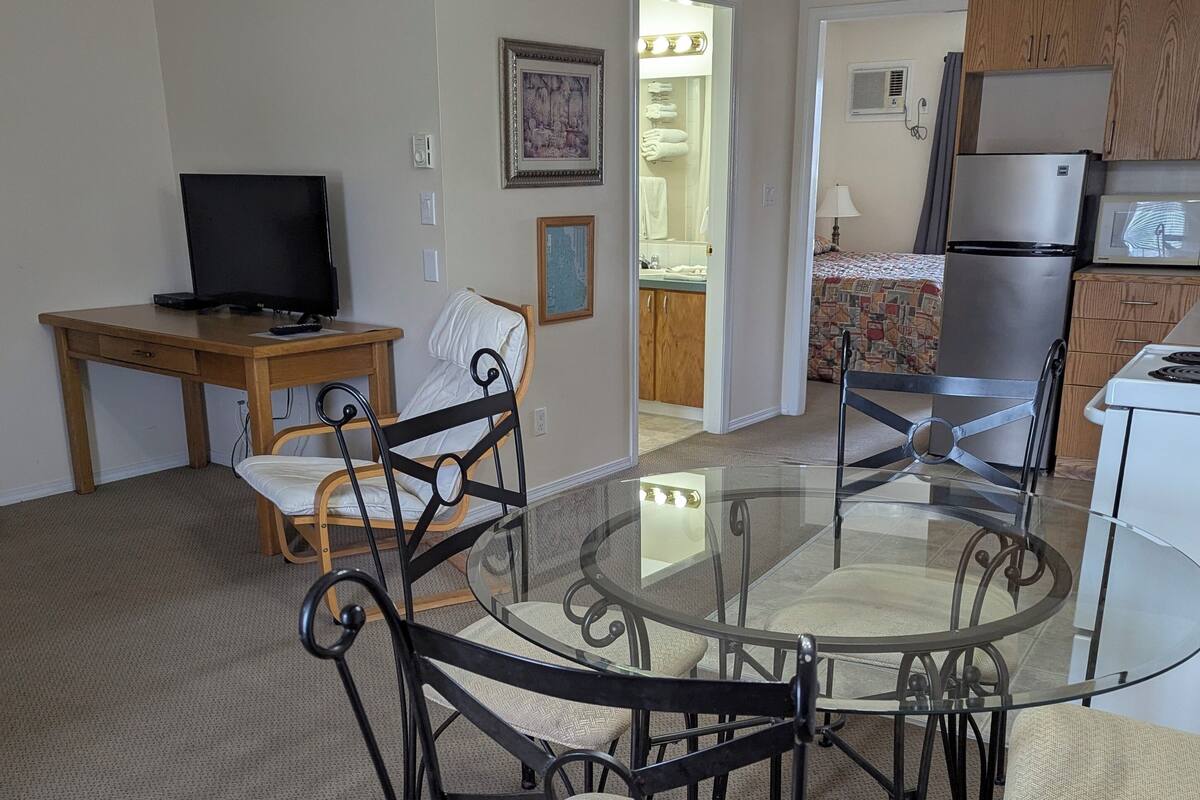 A living room with a glass-top dining table, black metal chairs, a wooden desk with a television, and a kitchen area in the background.