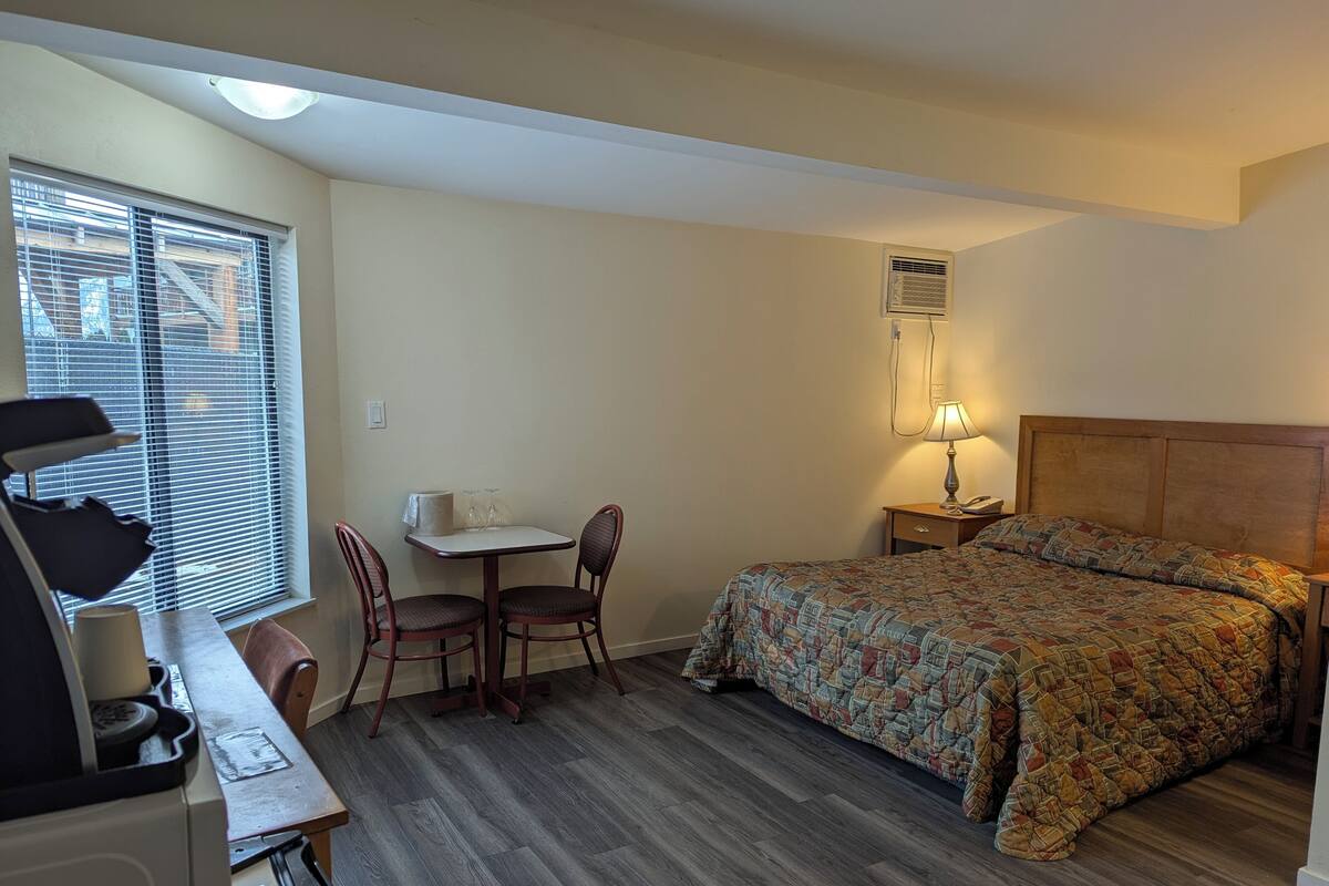A compact hotel room with a bed, a desk with a chair, a small table with a chair, a lamp, and a window with blinds.