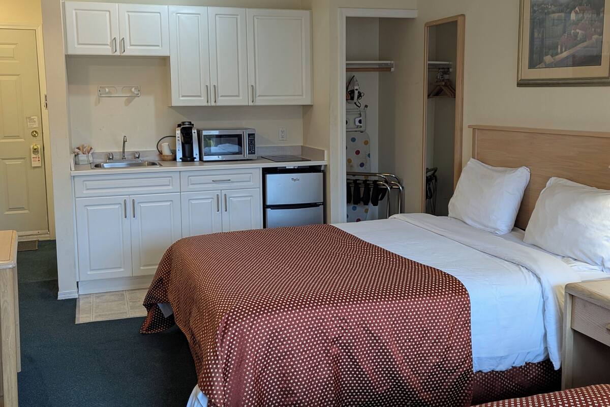 A compact hotel room with a kitchenette, a bed with a patterned bedspread, and a painting on the wall.