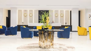 Un vestíbulo moderno con una mesa redonda central, asientos en tonos azul y amarillo, y elementos decorativos como relojes y plantas.