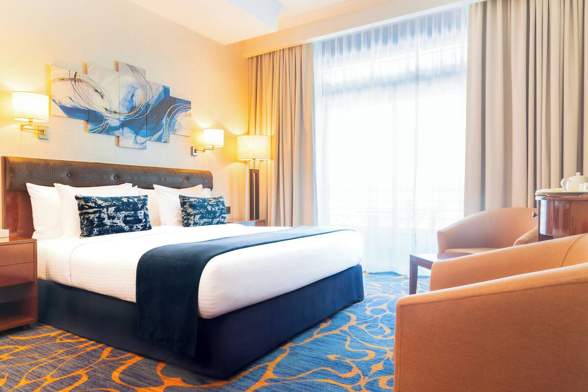 Habitación de hotel con una cama grande, dos lámparas de noche, cabecera con panel decorativo, un ventanal con cortinas y suelo cubierto de alfombra.